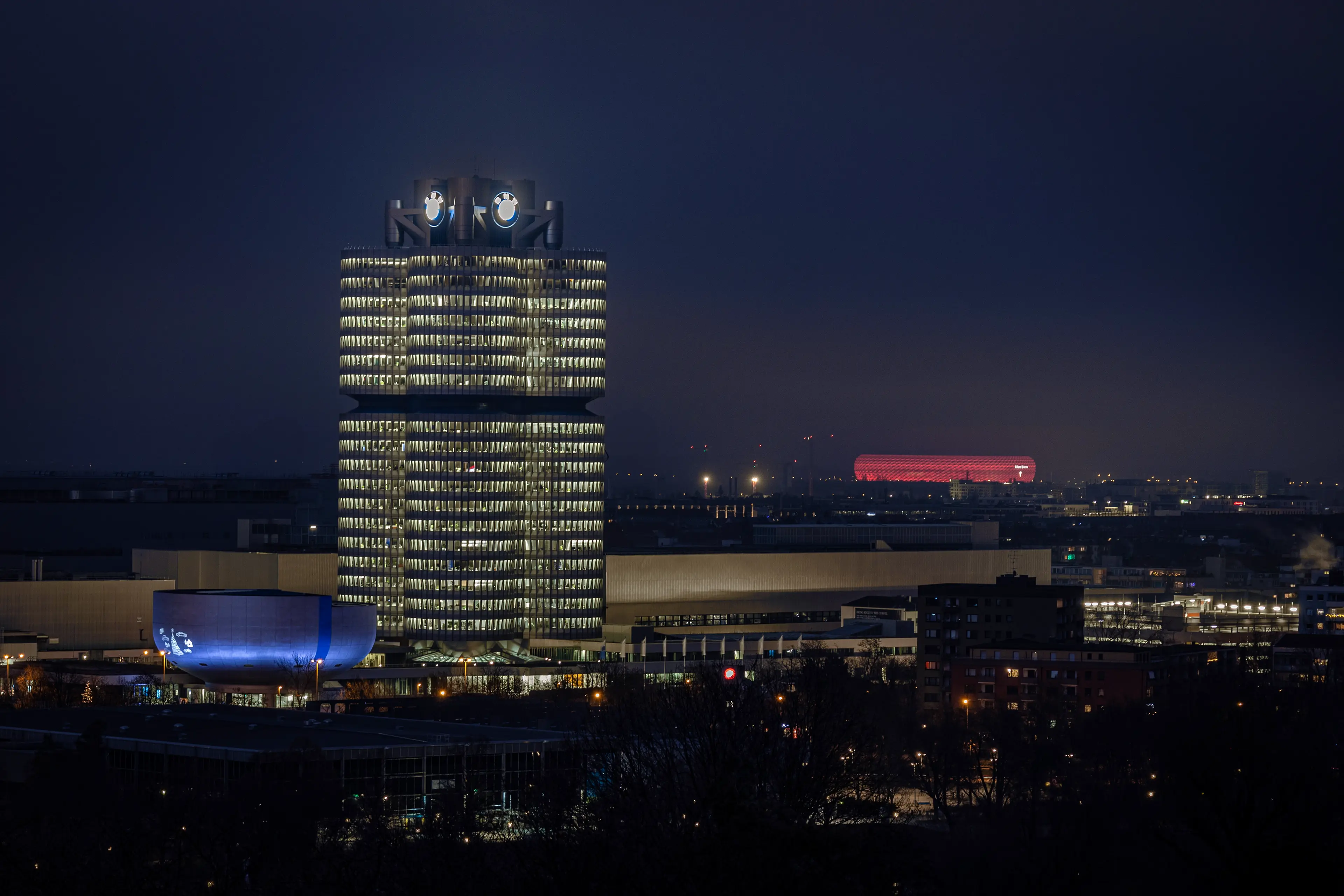 Das beleuchtete BMW-Vierzylinder-Hauptquartier in Muenchen bei Nacht, mit der rot leuchtenden Allianz Arena im Hintergrund.