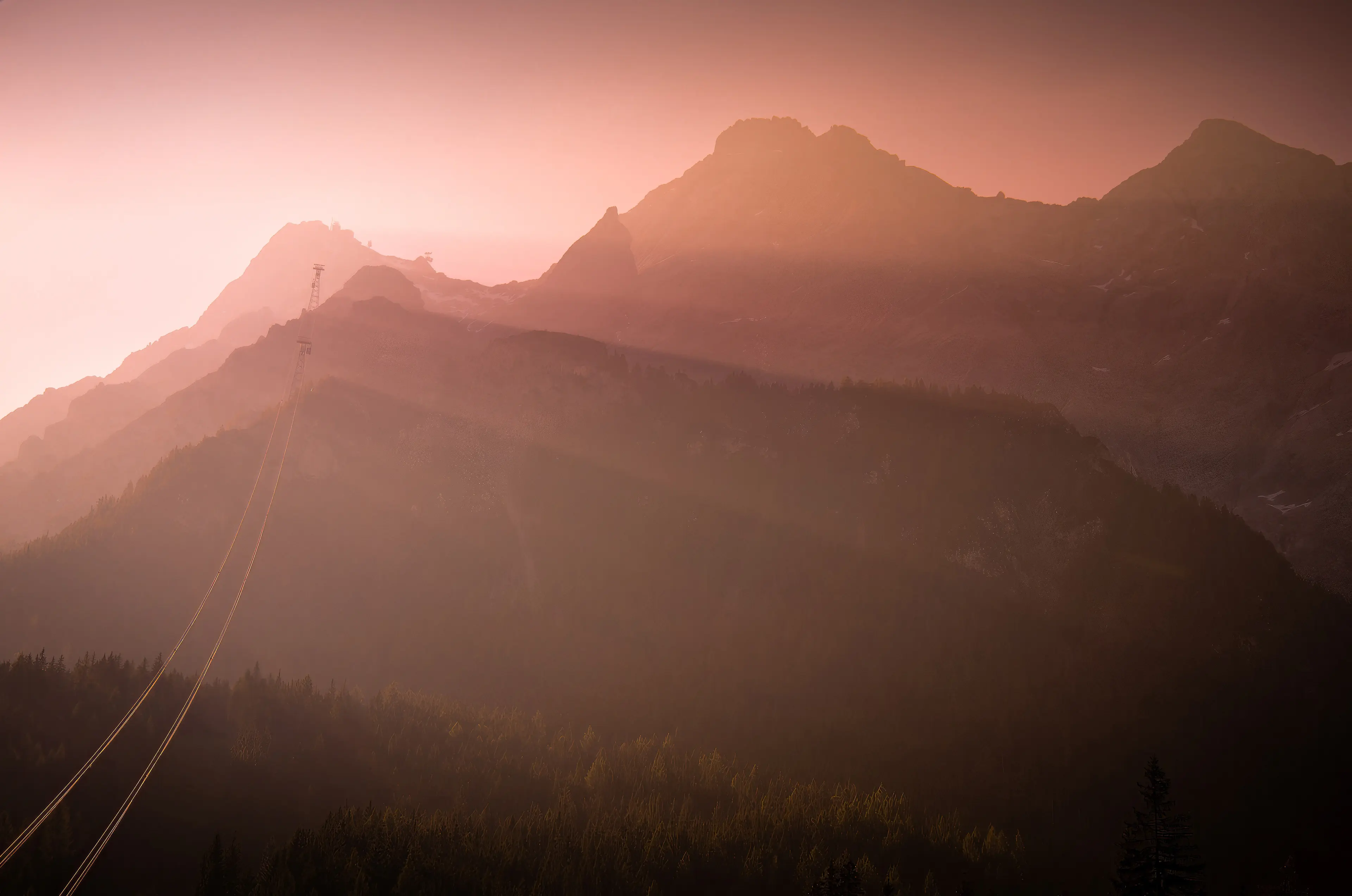 Silhouetten von Berggipfeln im ersten Morgenlicht mit einer Seilbahn im Dunst.