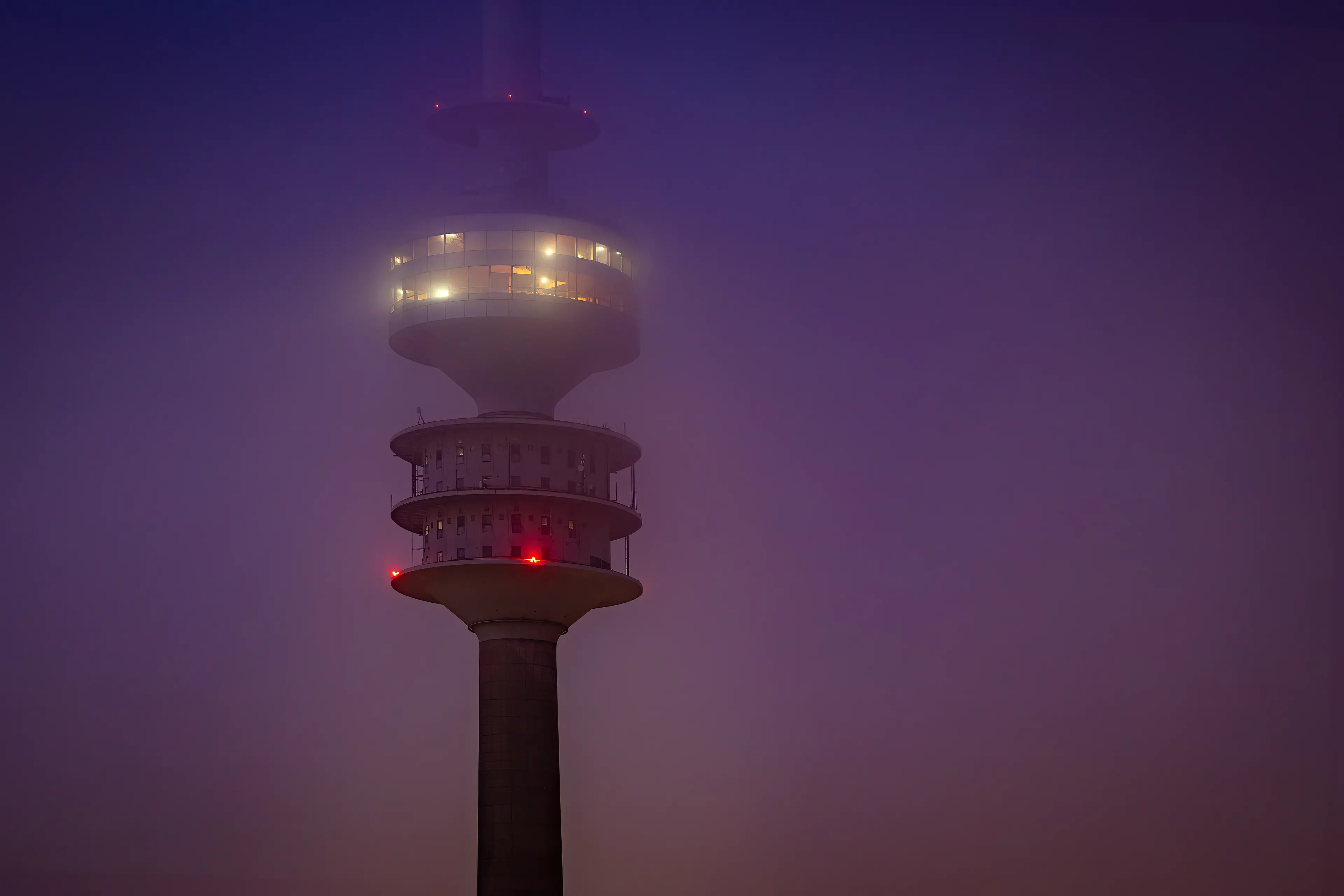 Der beleuchtete Münchner Olympiaturm, dessen obere Ebenen mystisch von dichtem Nebel in der abendlichen Dämmerung umhüllt sind.