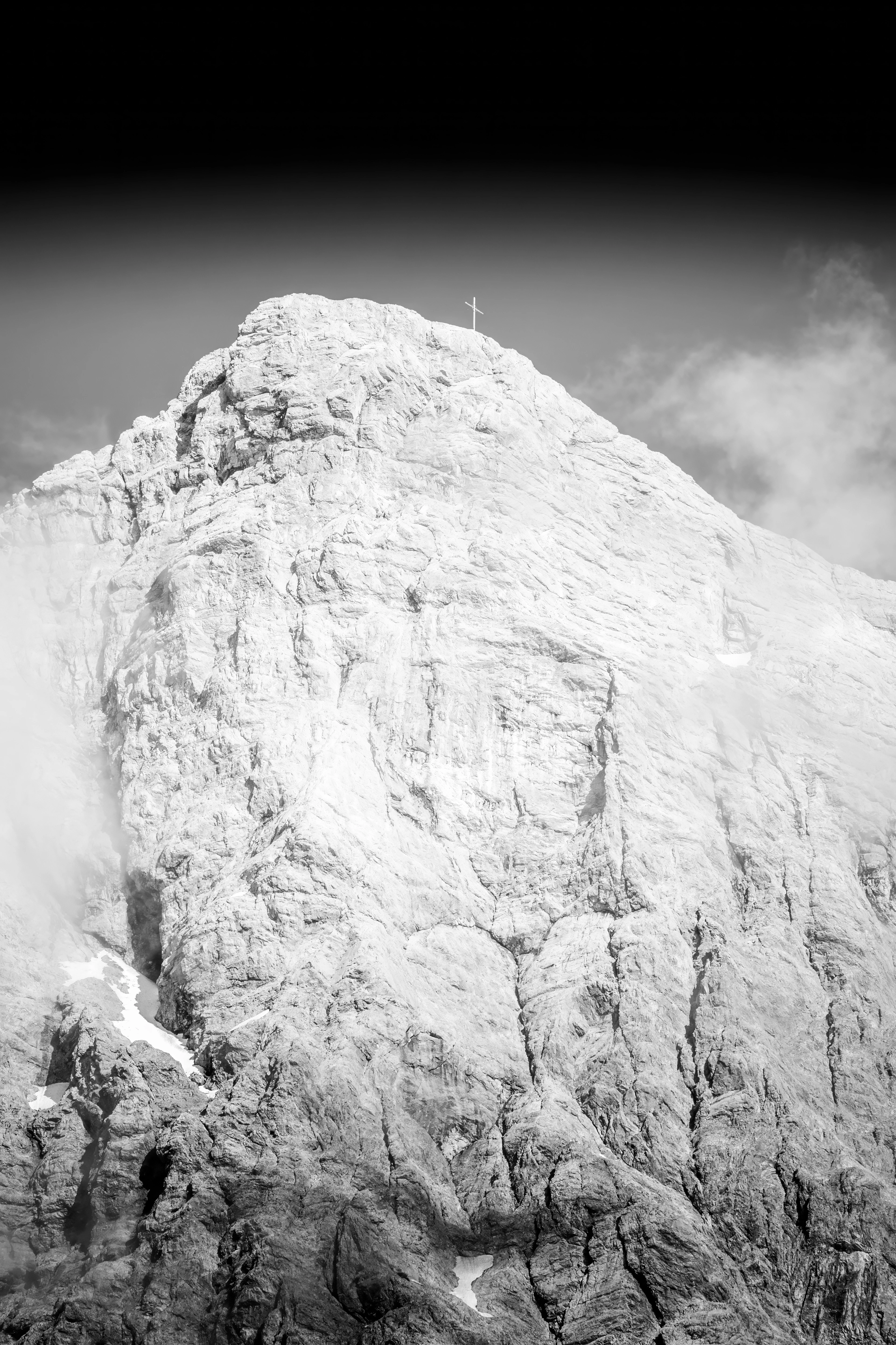 Schwarz-weiße Nahaufnahme eines schroffen Berggipfels mit Gipfelkreuz vor dunklem Himmel.
