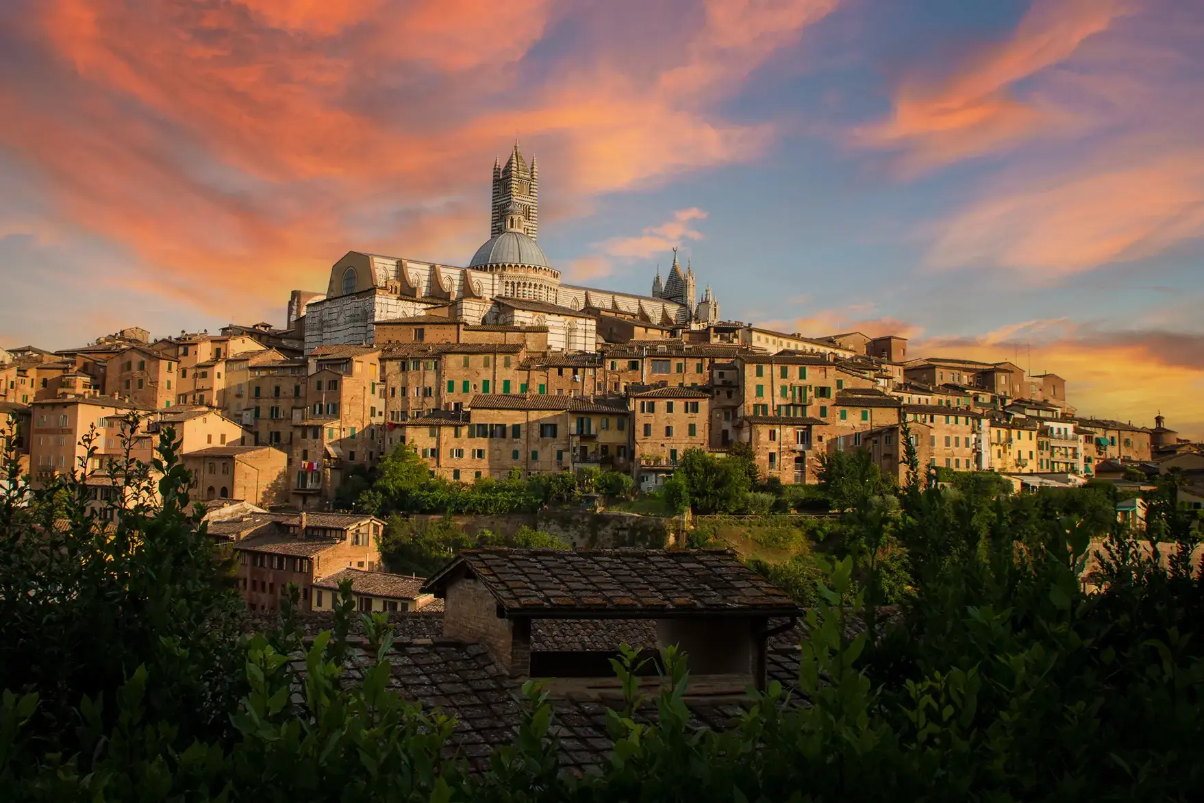 Historische Altstadt von Siena mit dem markanten Dom, eingebettet in eine Landschaft unter einem farbenfrohen Sonnenuntergangshimmel.