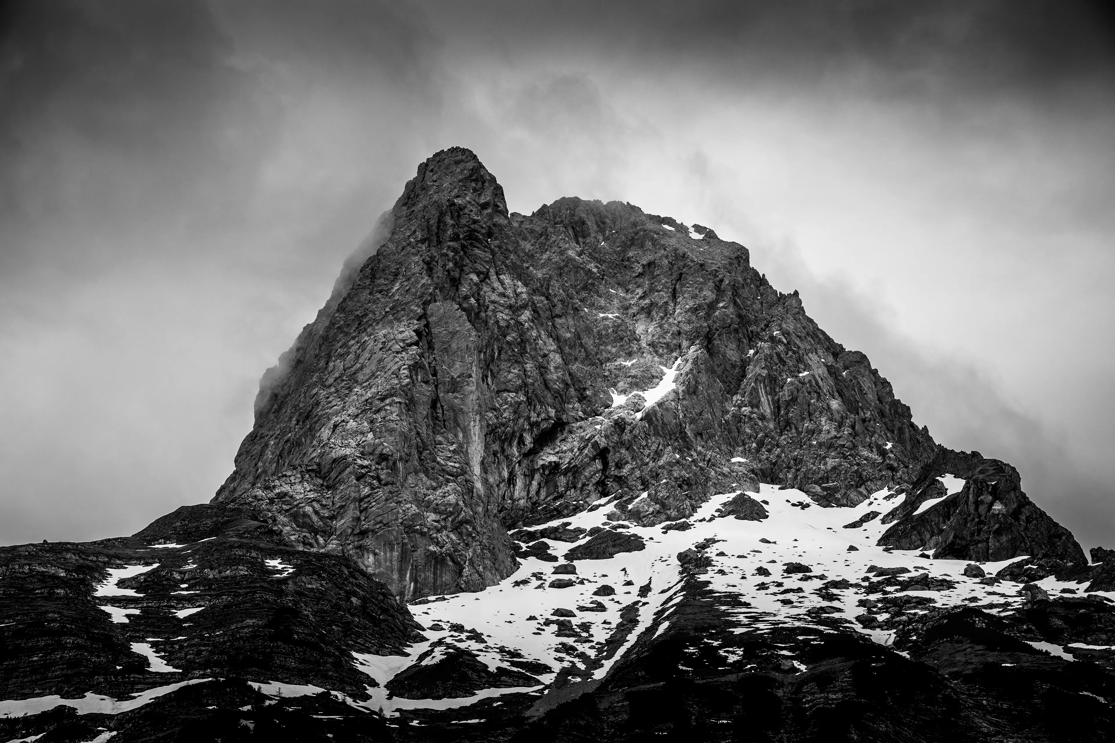 Kontrastreiche Schwarz-Weiß-Aufnahme eines massiven, spitzen Berggipfels, an dessen Fuss noch Schneereste liegen.