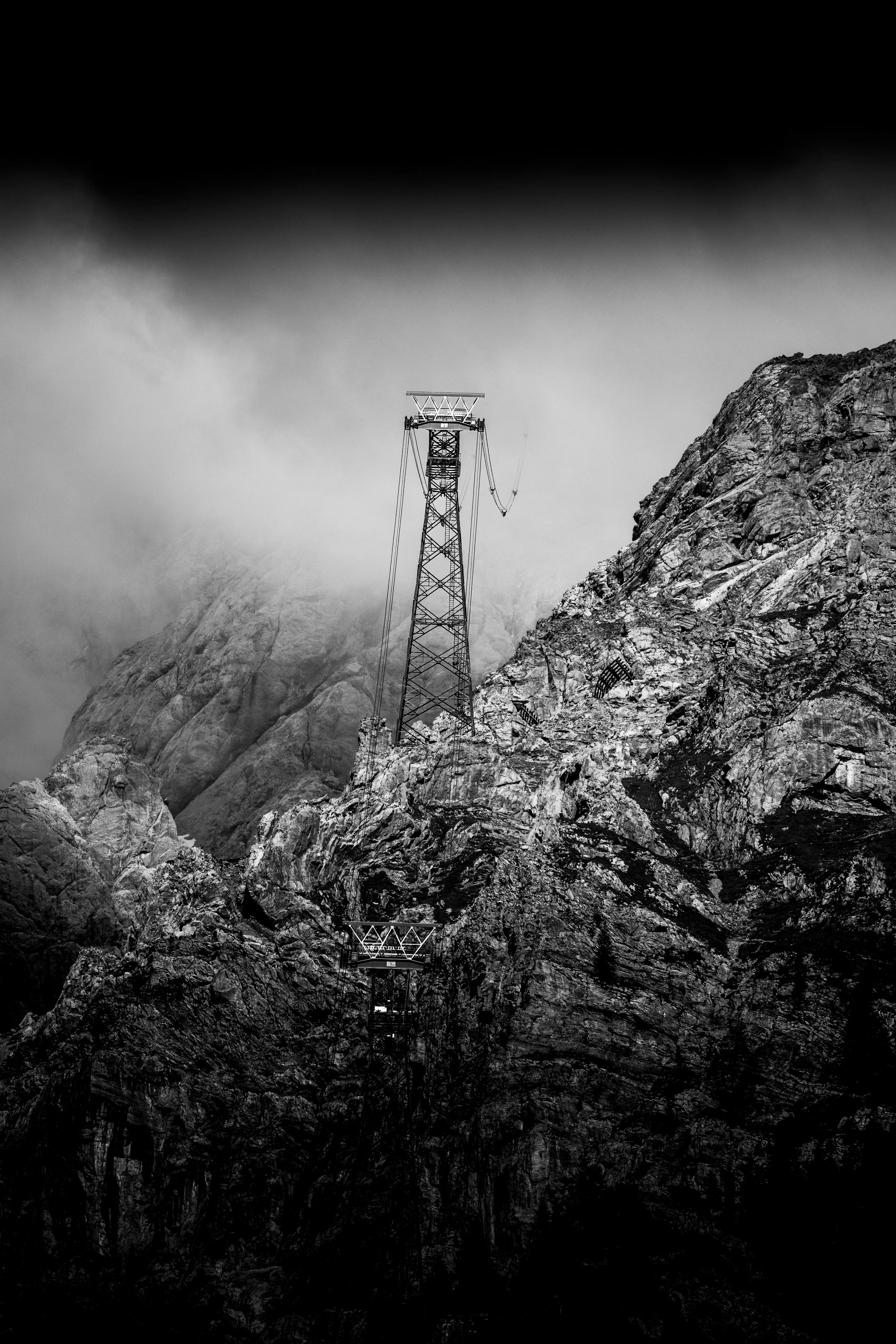 Schwarz-Weiss-Fotografie einer steilen Felswand, an der ein grosser, markanter Stahlmast einer Seilbahn thront.