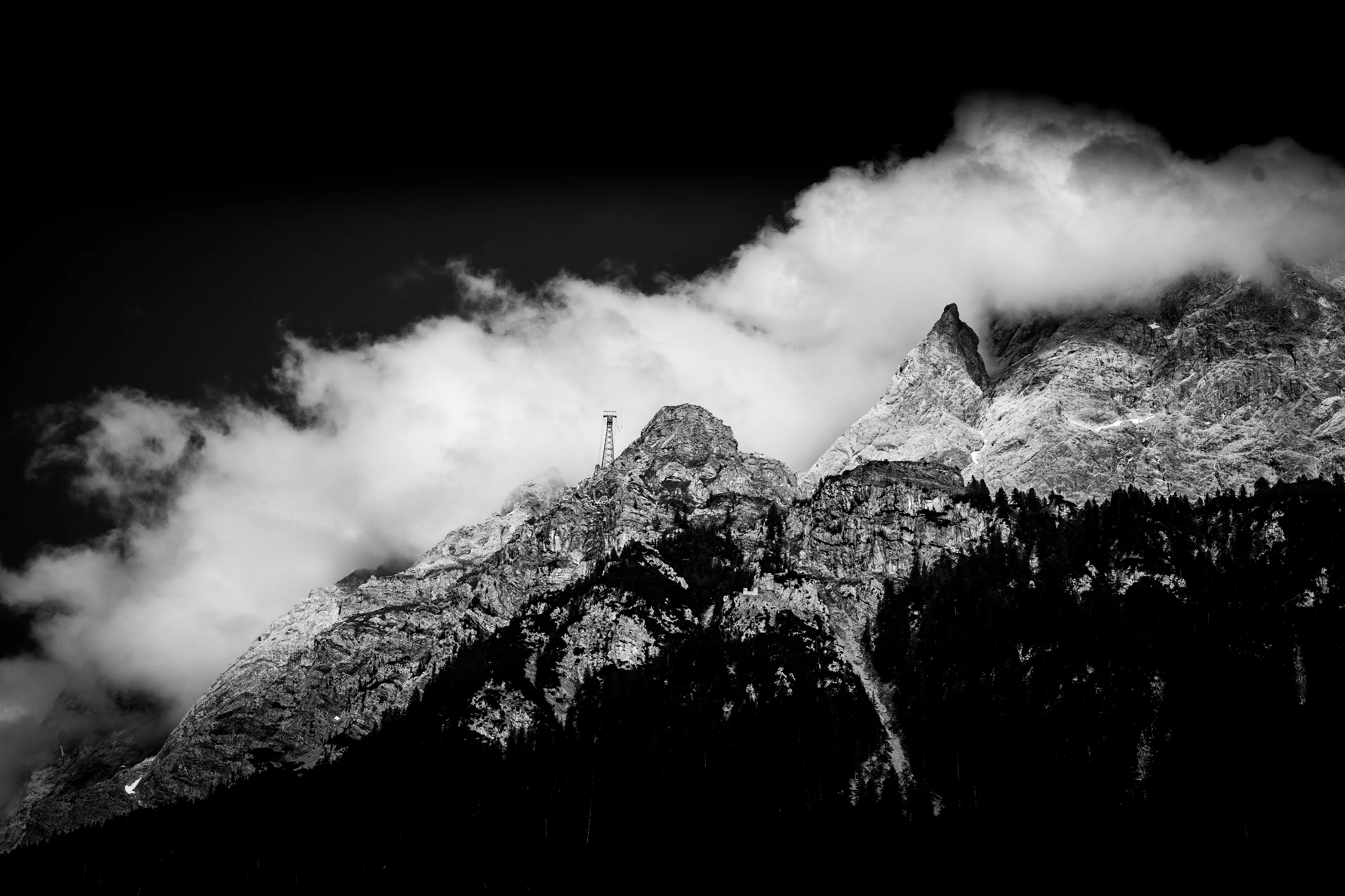 Dramatische Schwarz-Weiß-Aufnahme eines schroffen, wolkenverhangenen Berggipfels mit einem sichtbaren Seilbahnmast auf dem Grat.